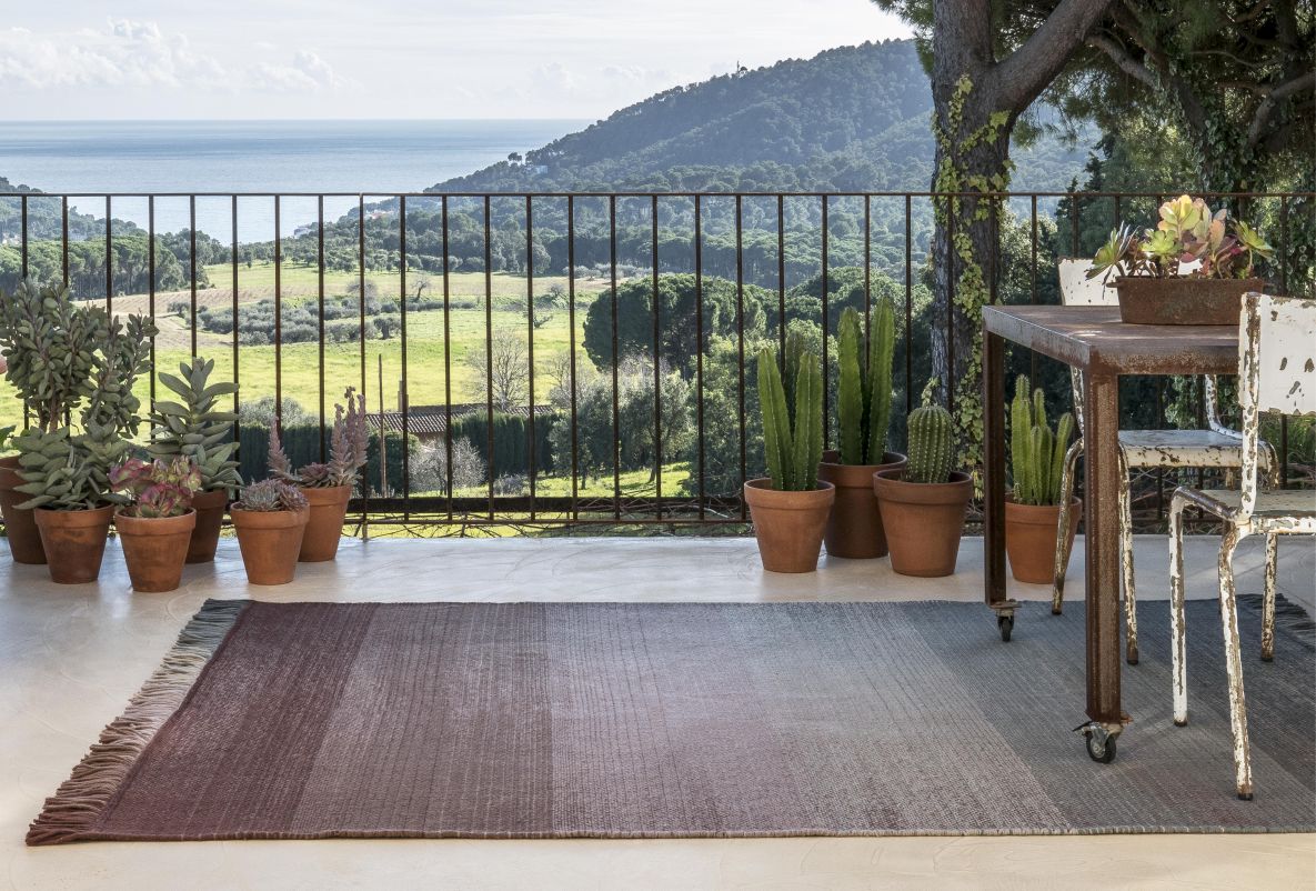 Handgewebter Teppich mit Farbverlauf in Braun-Grau auf einer Terrasse mit Meerblick und Pflanzen.