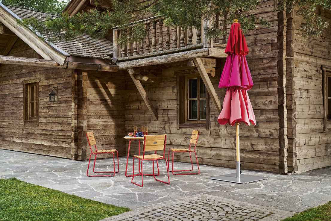 Weishäupl Klassiker Sonnenschirm in Rosa- und Rottönen neben rustikaler Holzhütte mit Gartenmöbeln für entspannte Stunden im Freien.