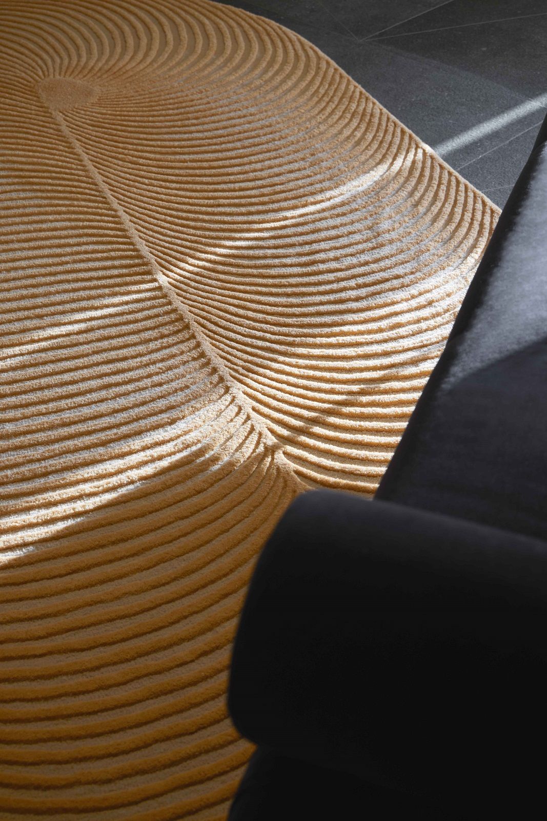 Detailaufnahme des beigen Giro Teppichs mit wellenförmigem Muster und schwarzem Sessel im Hintergrund.
