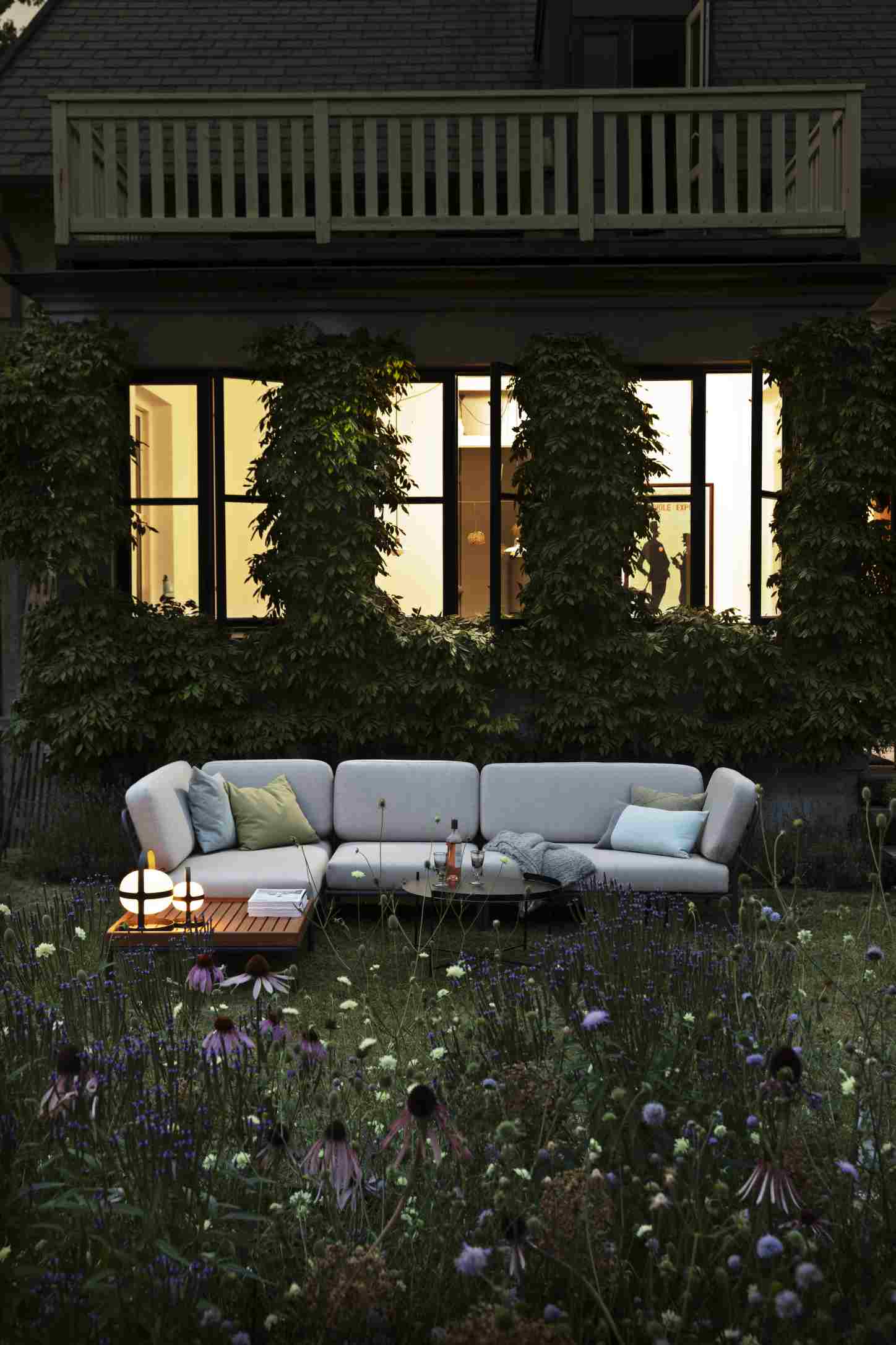 Houe Level Outdoor Sofa auf einer Terrasse mit Garten und blühenden Blumen im Vordergrund.