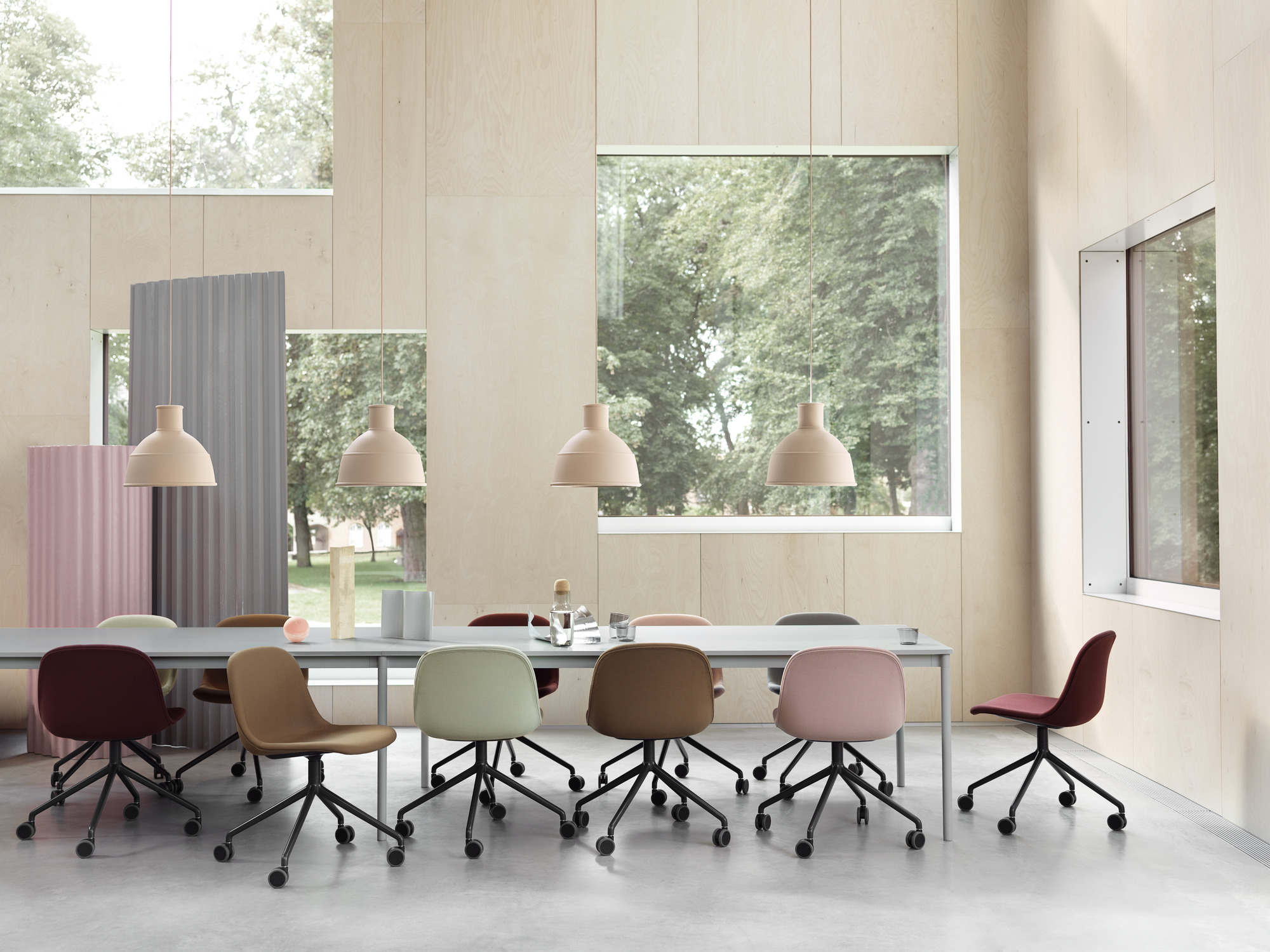 Helles Büro mit langem Tisch und farbigen Stühlen, darüber hängen Lampen. Muuto Tisch und Stühle.