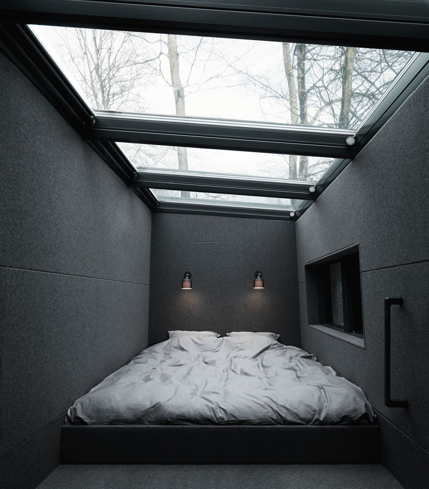 Schlafzimmer mit Bett und zwei schwarzen Wandspots von Vipp unter einem Glasdach mit Blick auf Bäume.