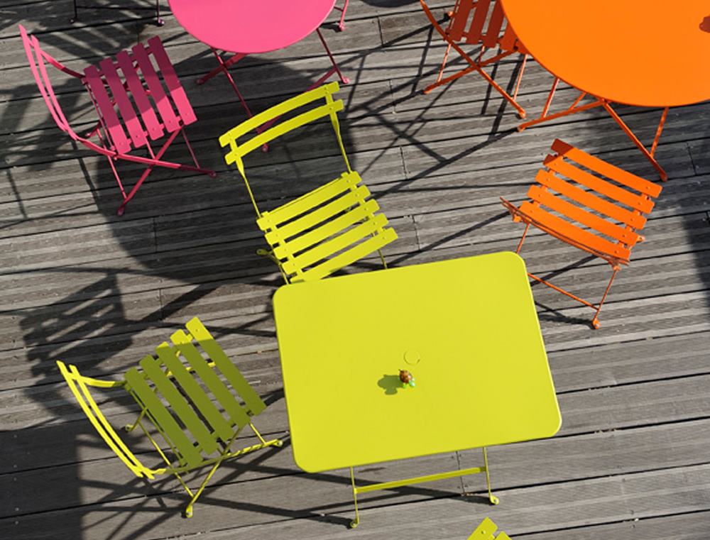 Draufsicht auf farbenfrohe Fermob Bistro Gartenmöbel: Klapptische und Stühle in Pink, Gelb und Orange auf Holzterrasse.