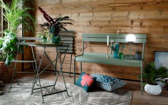 Grüner Bistro+ Tisch von Fermob mit Stühlen und Bank im Outdoor-Bereich vor Holzwand. Gartenmöbel für Balkon und Terrasse.