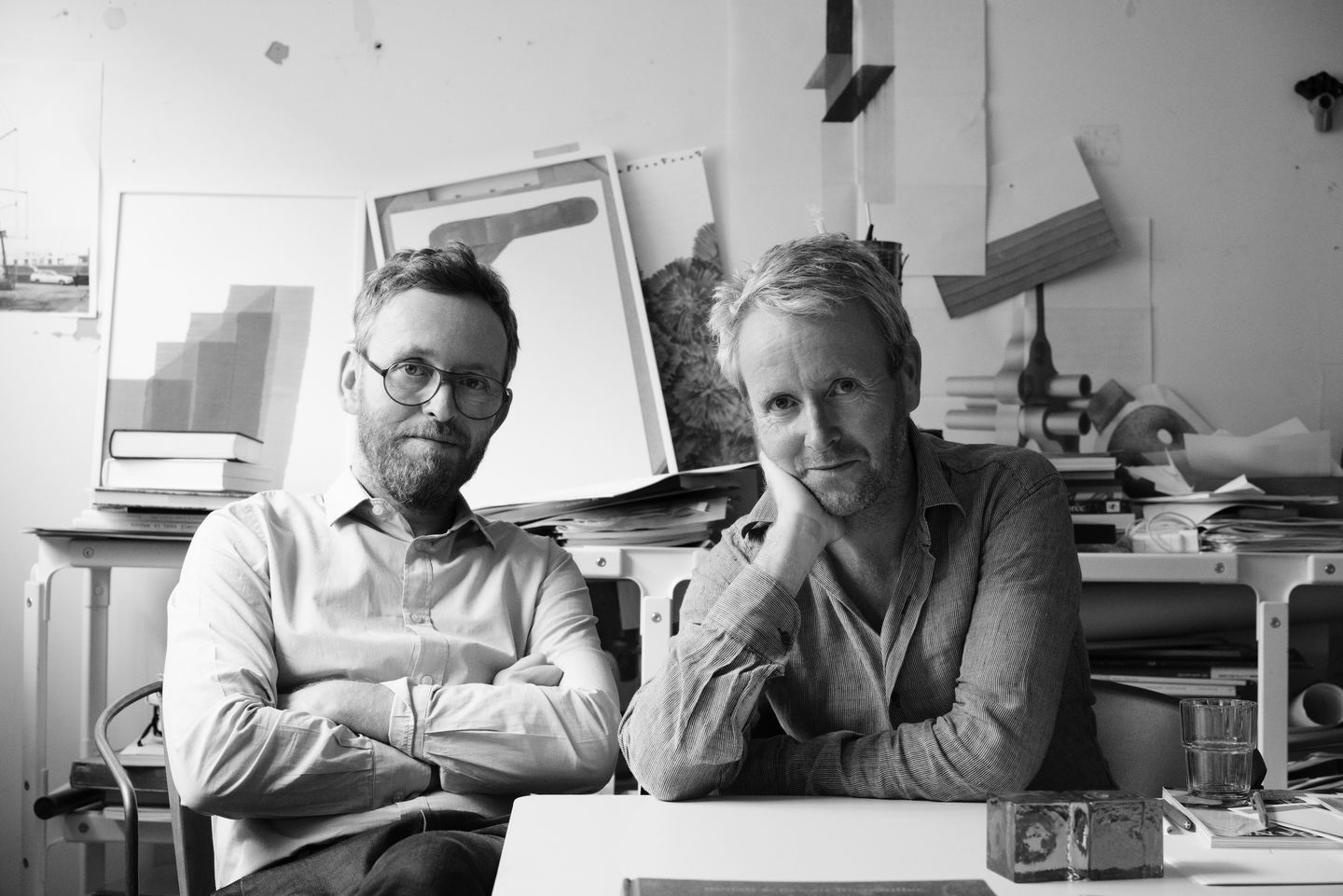 Schwarzweiß-Porträt zweier Künstler in ihrem Atelier, umgeben von Kunstwerken und Arbeitsmaterialien.