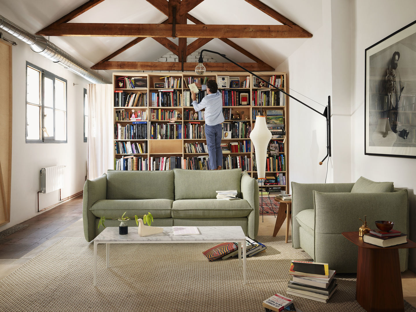Helles Wohnzimmer mit grünem Sofa, Sessel, Bücherregal und Person, die ein Buch entnimmt.