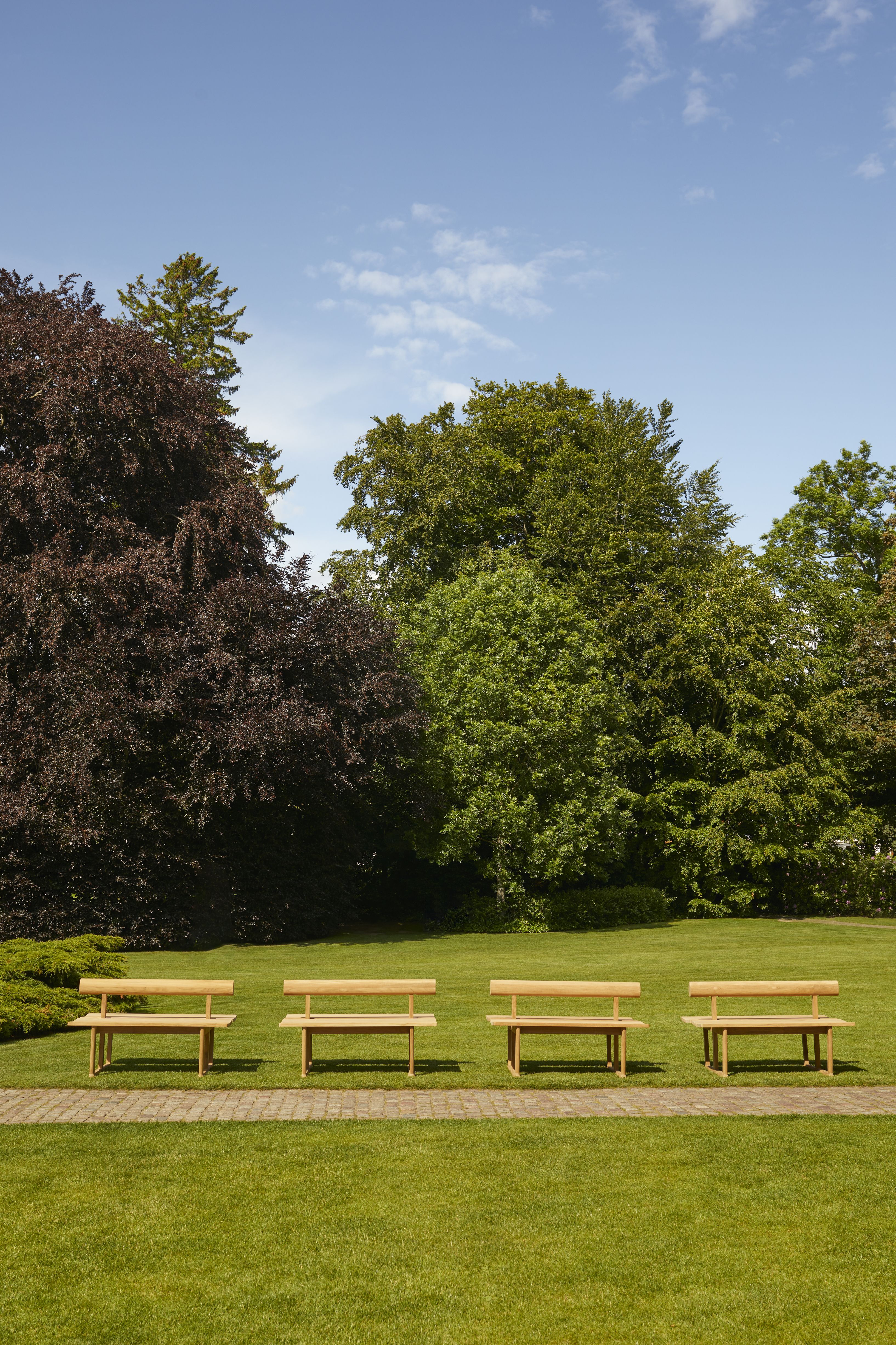 Vier Skagerak Banco Teak Gartenbänke auf Rasen vor Bäumen unter blauem Himmel, Outdoor Möbel.