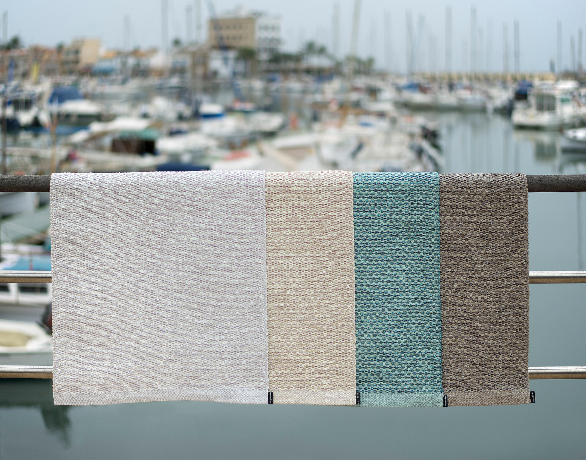 Svea Pappelina Kunststoffteppich in Weiß, Beige, Türkis und Braun vor einem Hafen mit Booten.