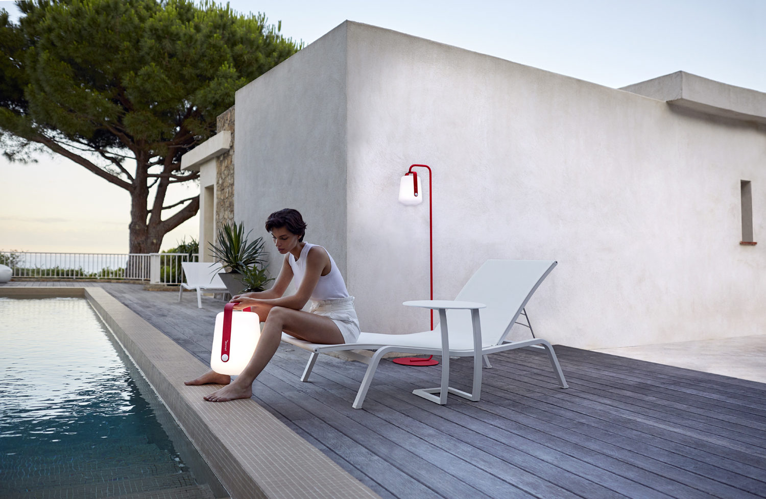 Frau sitzt am Pool mit Alizé Outdoor Tischleuchte und roter Stehlampe von Fermob im modernen Garten.