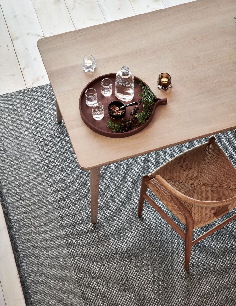 Grauer Edit Teppich von Pappelina (230x320cm) unter einem Holztisch mit Dekoration und Stuhl.