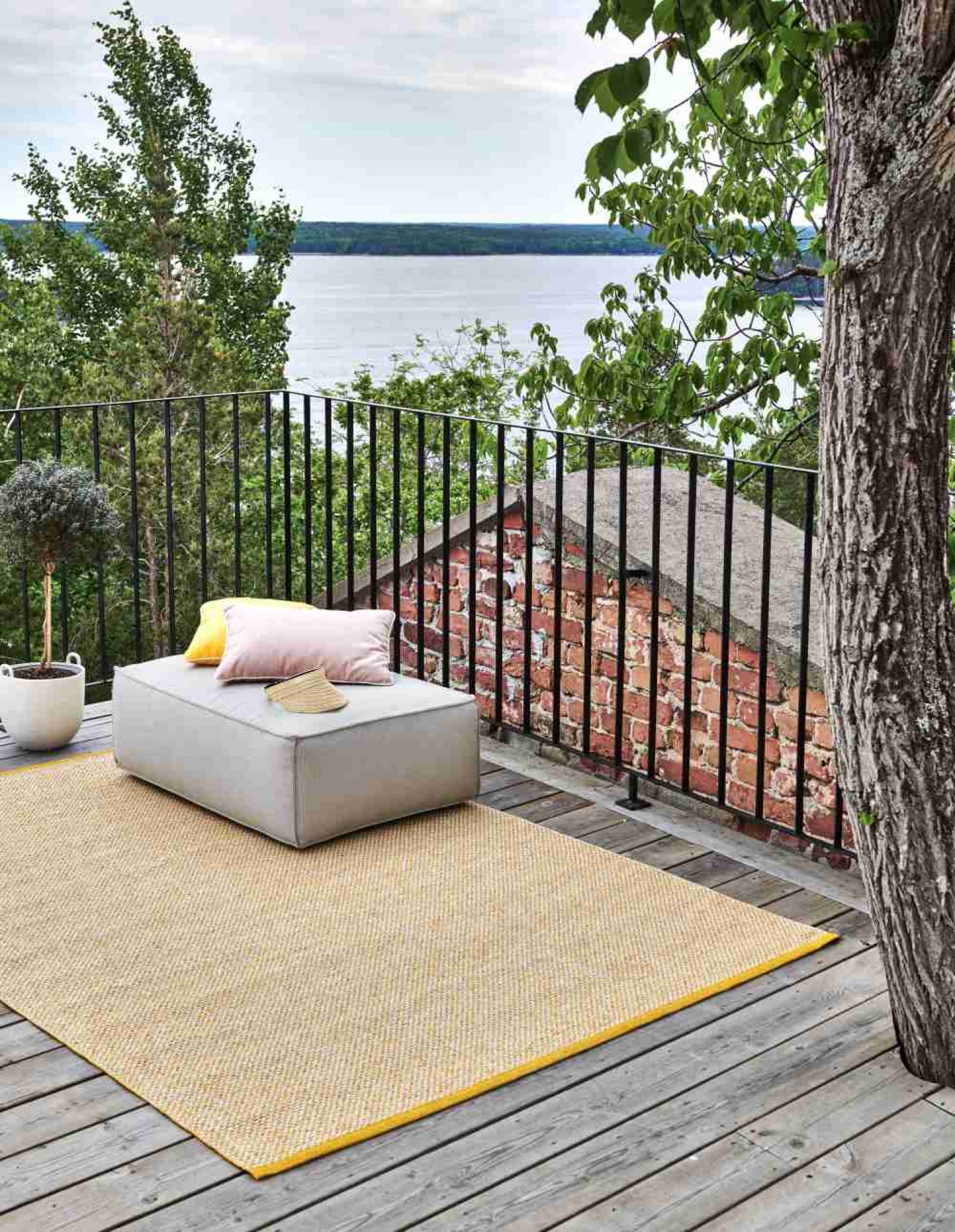 Gelber Outdoor Teppich auf Holzterrasse mit Hocker, Kissen und Blick auf den See.
