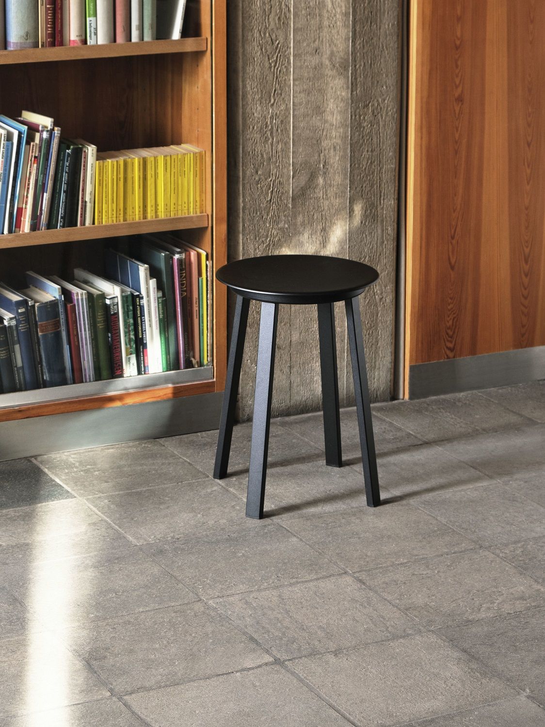 Schwarzer Revolver Stool Hocker von Hay vor Bücherregal und Holzwand, modernes Möbeldesign für Wohnraum.