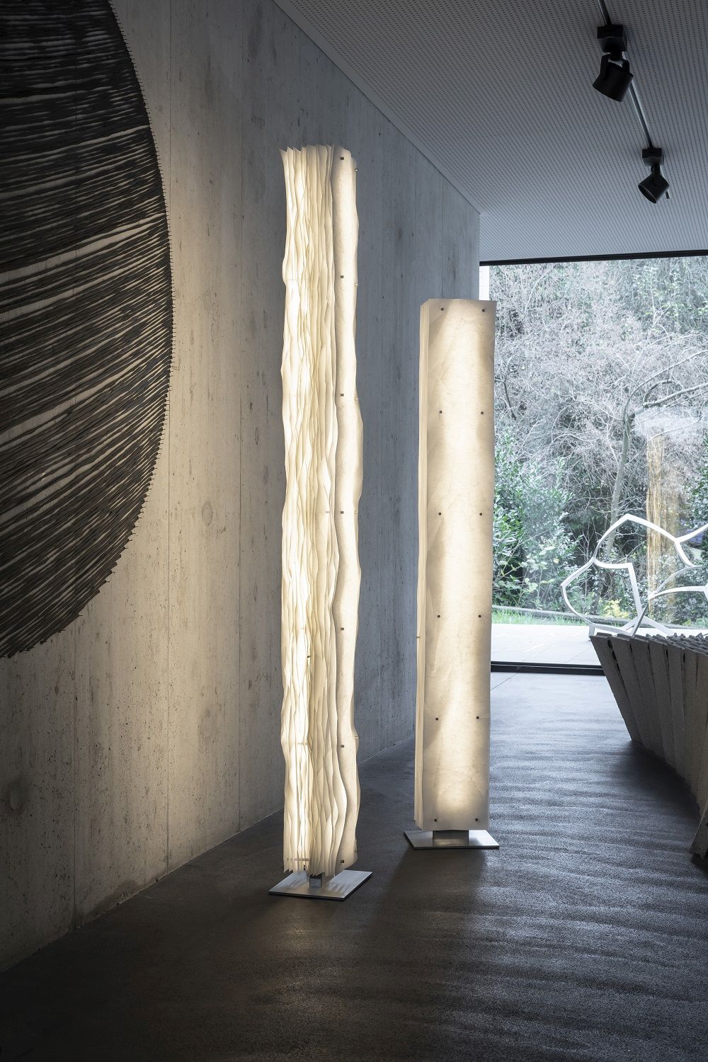 Zwei moderne Totem Stehleuchten im Stele Design, beleuchtet einen minimalistischen Flur mit Betonwand.