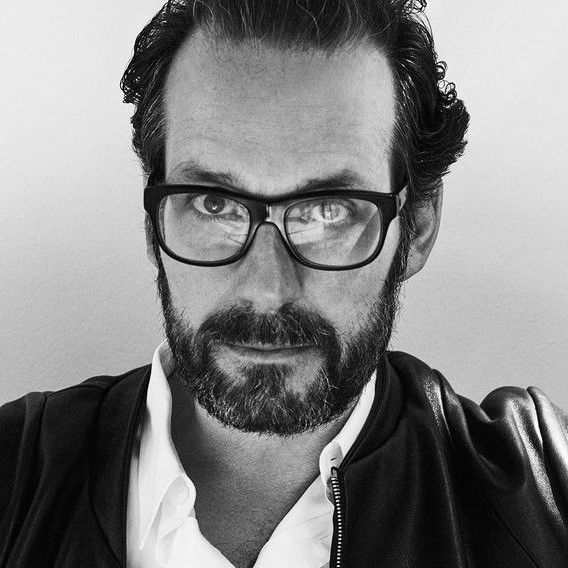Schwarz-Weiß-Porträt eines Mannes mit Bart und Brille, trägt eine Lederjacke und ein Hemd.