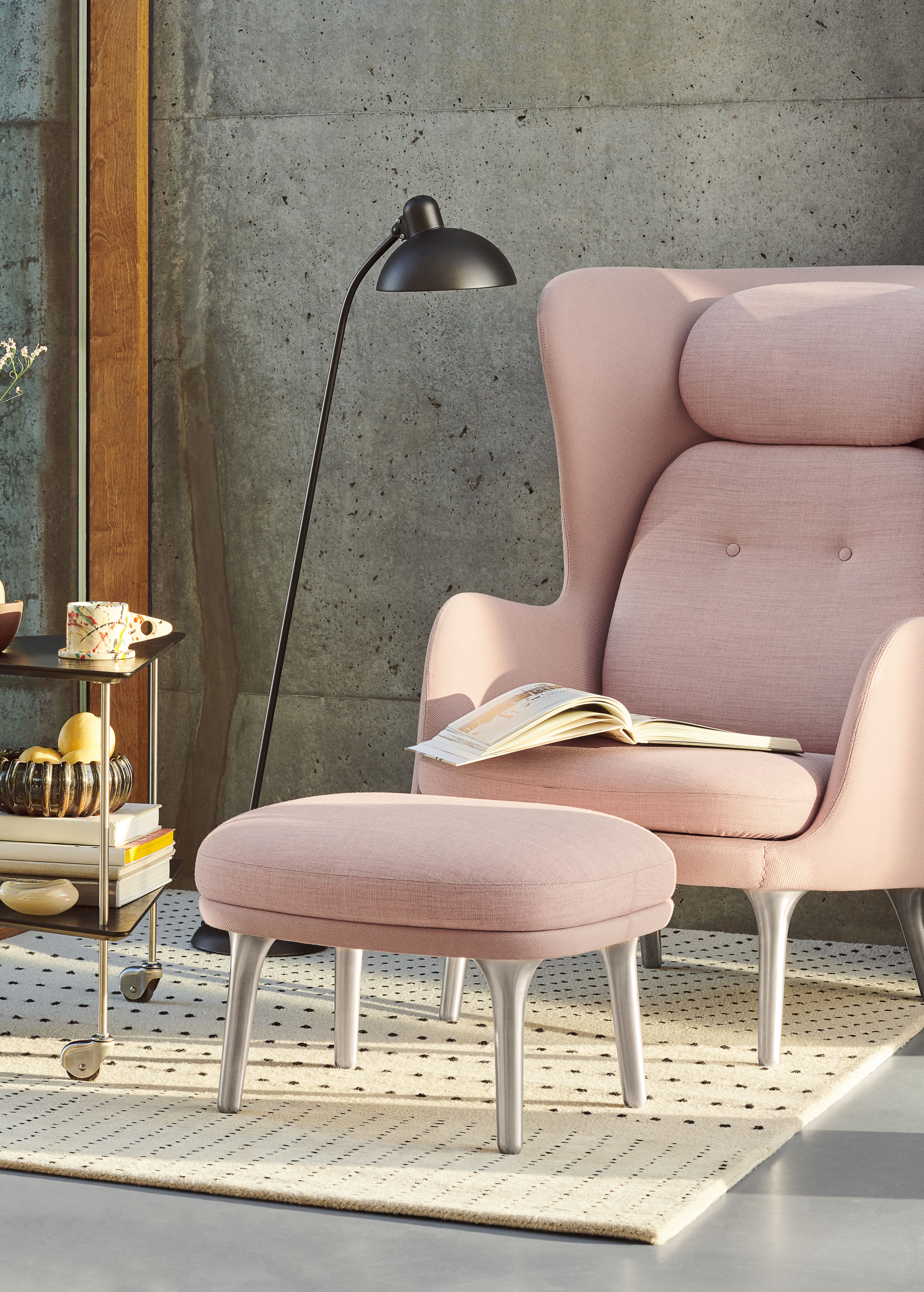 Rosa Fritz Hansen Ro Sessel mit Hocker und Buch auf Sitzfläche vor Betonwand.