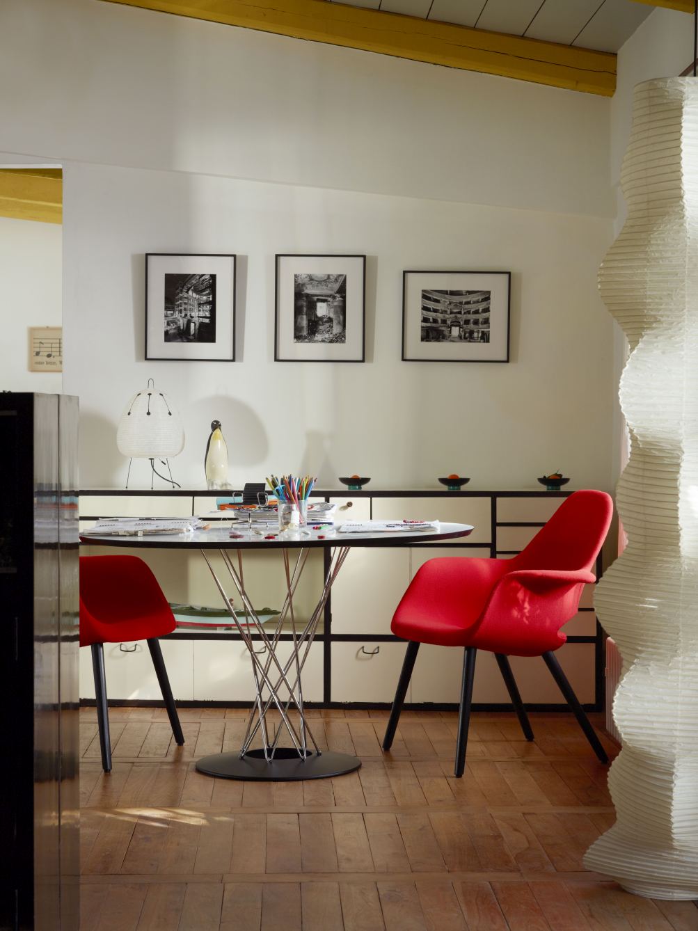 Vitra Dining Table Tisch Noguchi mit Glasplatte und verchromtem Stahlgestell, umgeben von roten Stühlen.