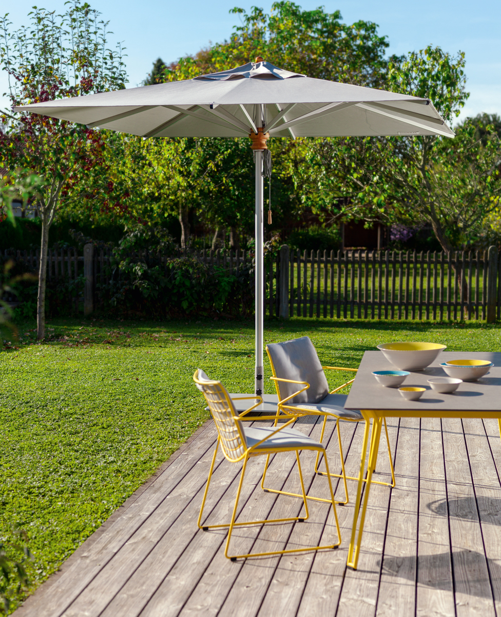 Grauer Alusonnenschirm von Weishäupl spendet Schatten über einer Gartenterrasse mit Tisch und Stühlen.