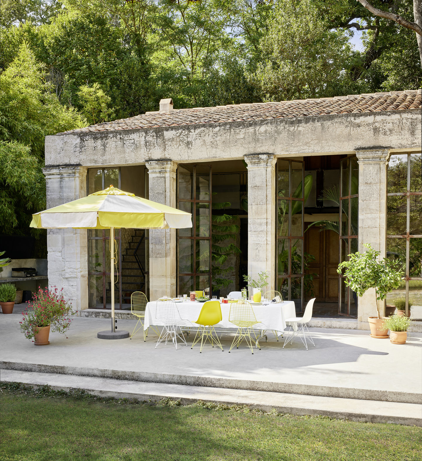 Gartenszene mit Vitra DKR Wire Chairs in Gelb und Weiß an gedecktem Tisch unter Sonnenschirm.