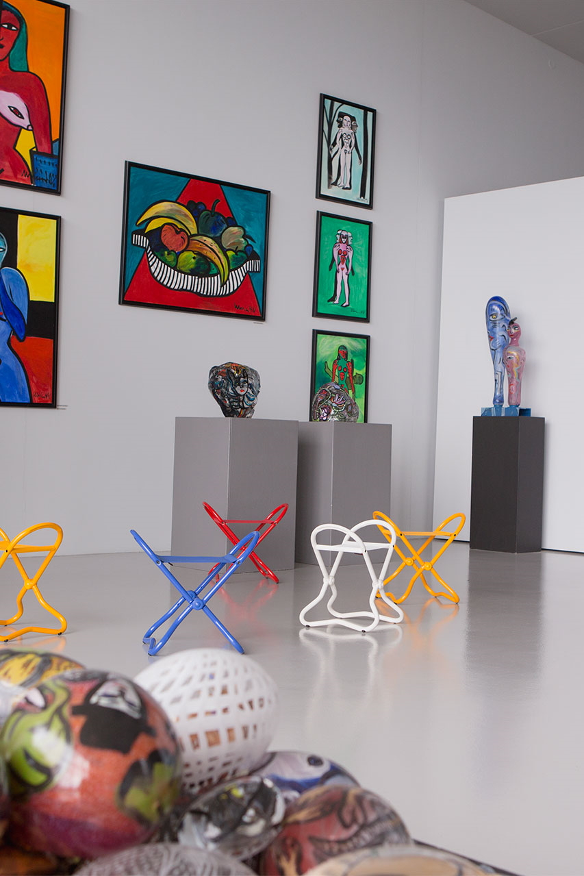 Ausstellung mit farbenfrohen Gemälden, Skulpturen und bunten Hockern in Gelb, Blau, Rot und Weiß.