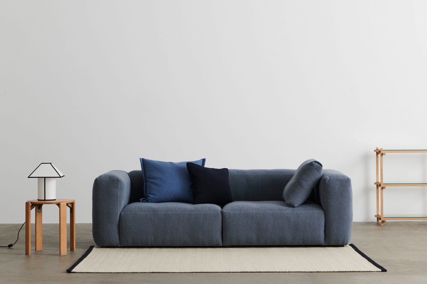 Blaues HAY Sofa mit Kissen, Lampe auf Holzhocker und Regal im minimalistischen Wohnzimmer.