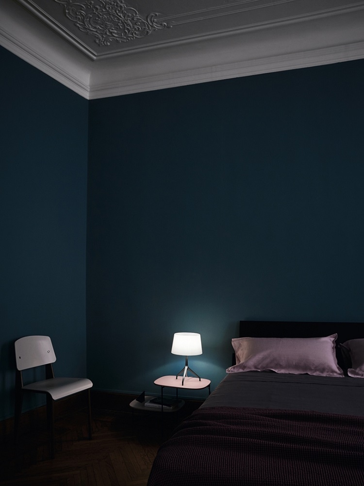 Schlafzimmer mit Lumiere Tischleuchte von Foscarini auf Nachttisch neben Bett und Stuhl.
