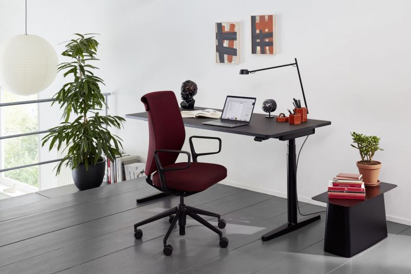 Ergonomischer Bürostuhl in Bordeauxrot vor schwarzem, höhenverstellbarem Schreibtisch mit Laptop und Büroutensilien.