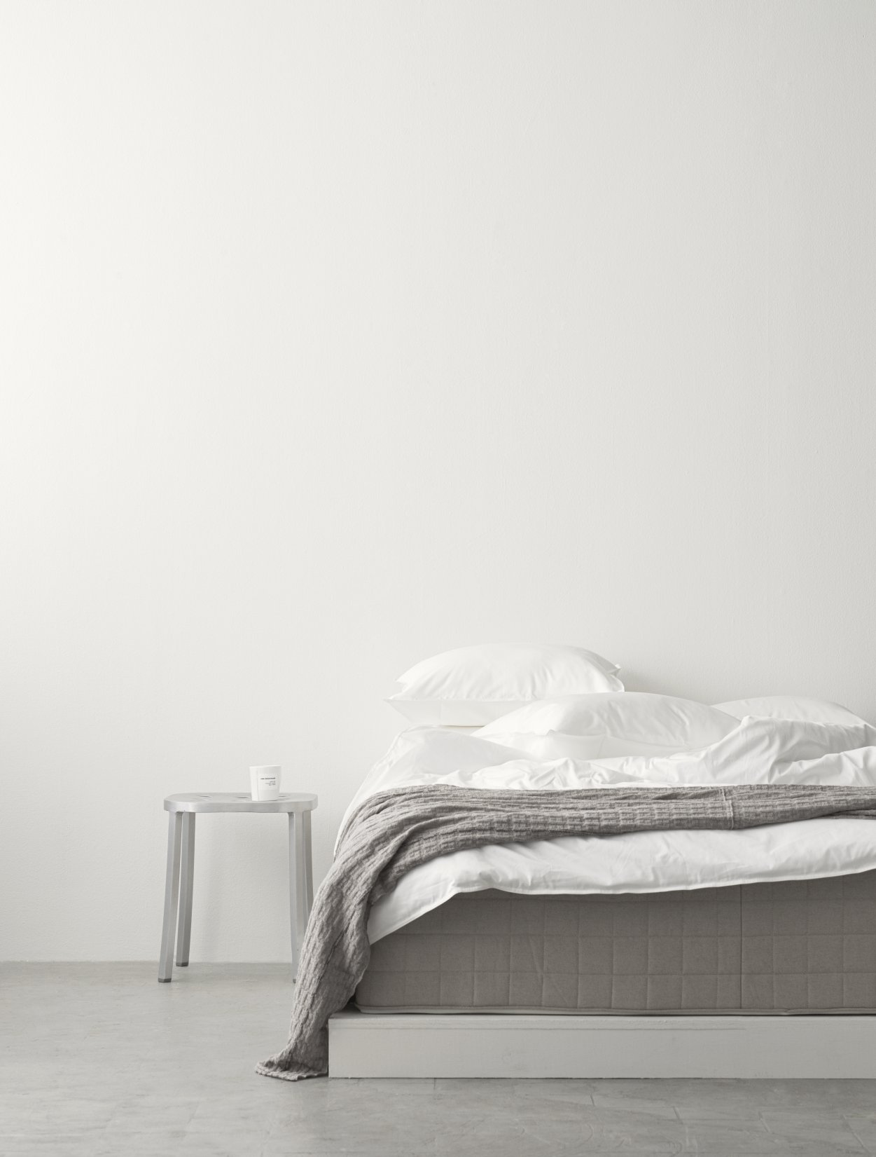 Foto eines 1 Inch Emeco Hockers aus Aluminium neben einem Bett mit Decke.