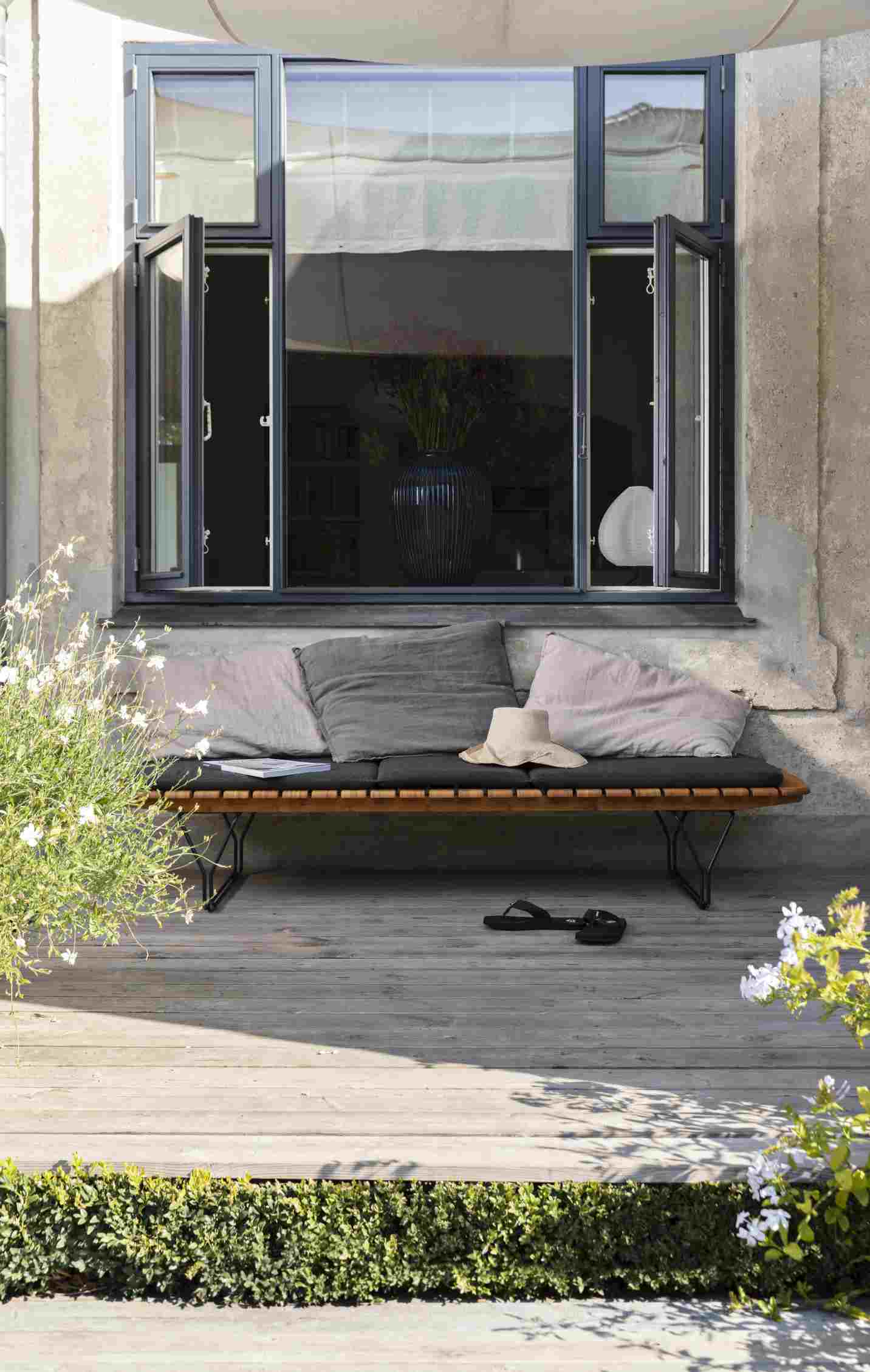 Houe Sonnenliege mit Molo Outdoor Kissen, grauer Bezug, hellrosa Zierkissen und Strohhut auf Terrasse.