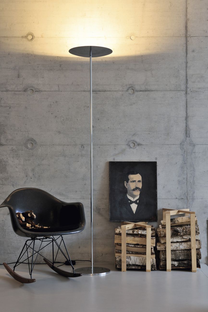 Moderne Stehlampe mit schwarzem Schirm, schwarzer Schaukelstuhl und Brennholz vor einer Betonwand.