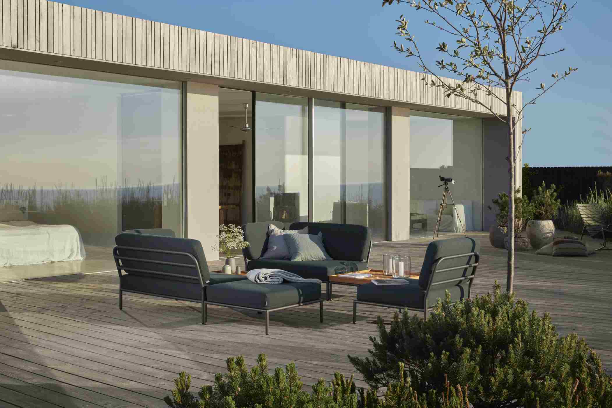 Houe Level Outdoor Sofa-Set auf Holzterrasse vor modernem Haus mit großen Glasfronten.