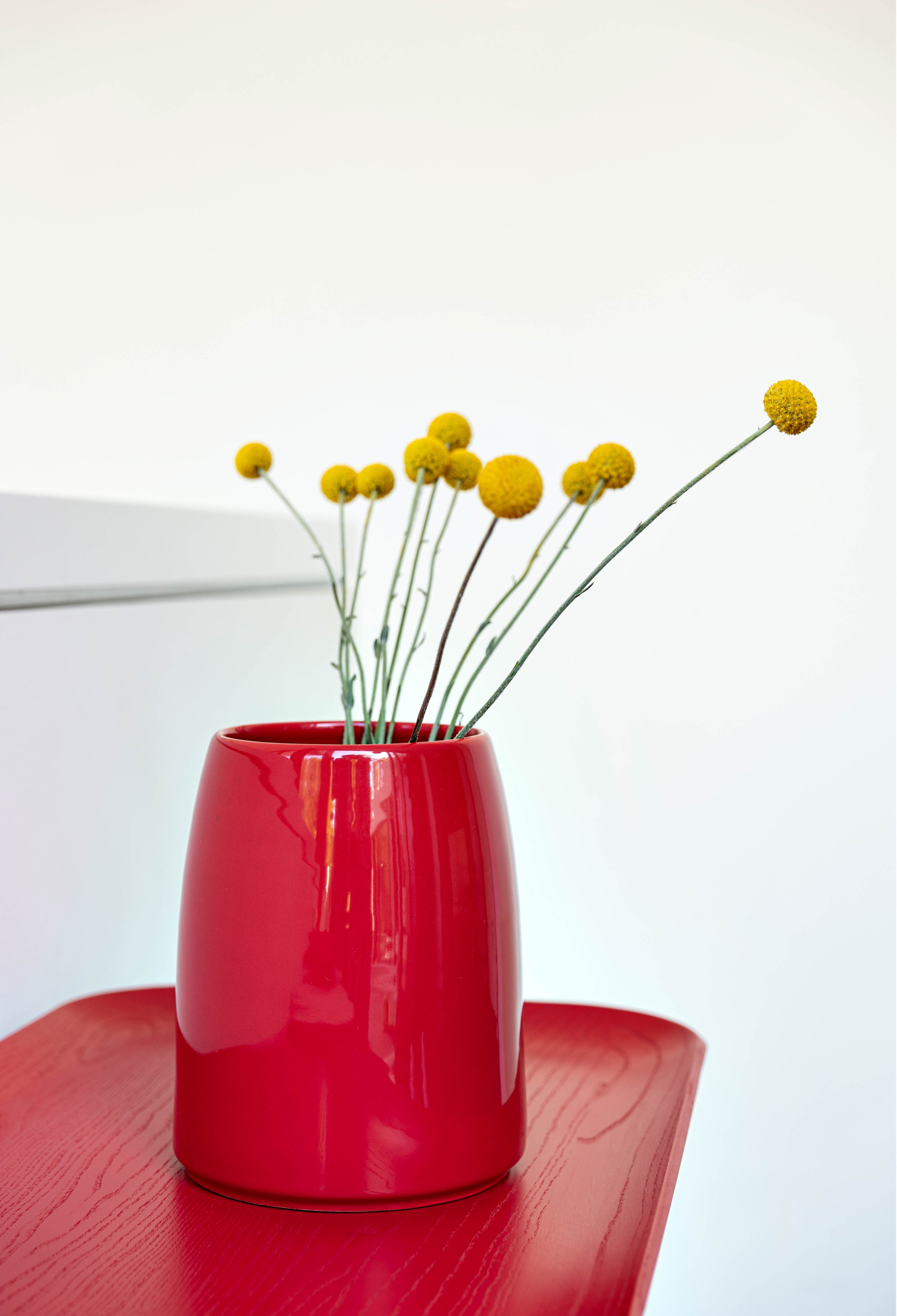 Rote Carla Vase von Schönbuch mit gelben Craspedia-Blüten auf einem passenden roten Tisch.