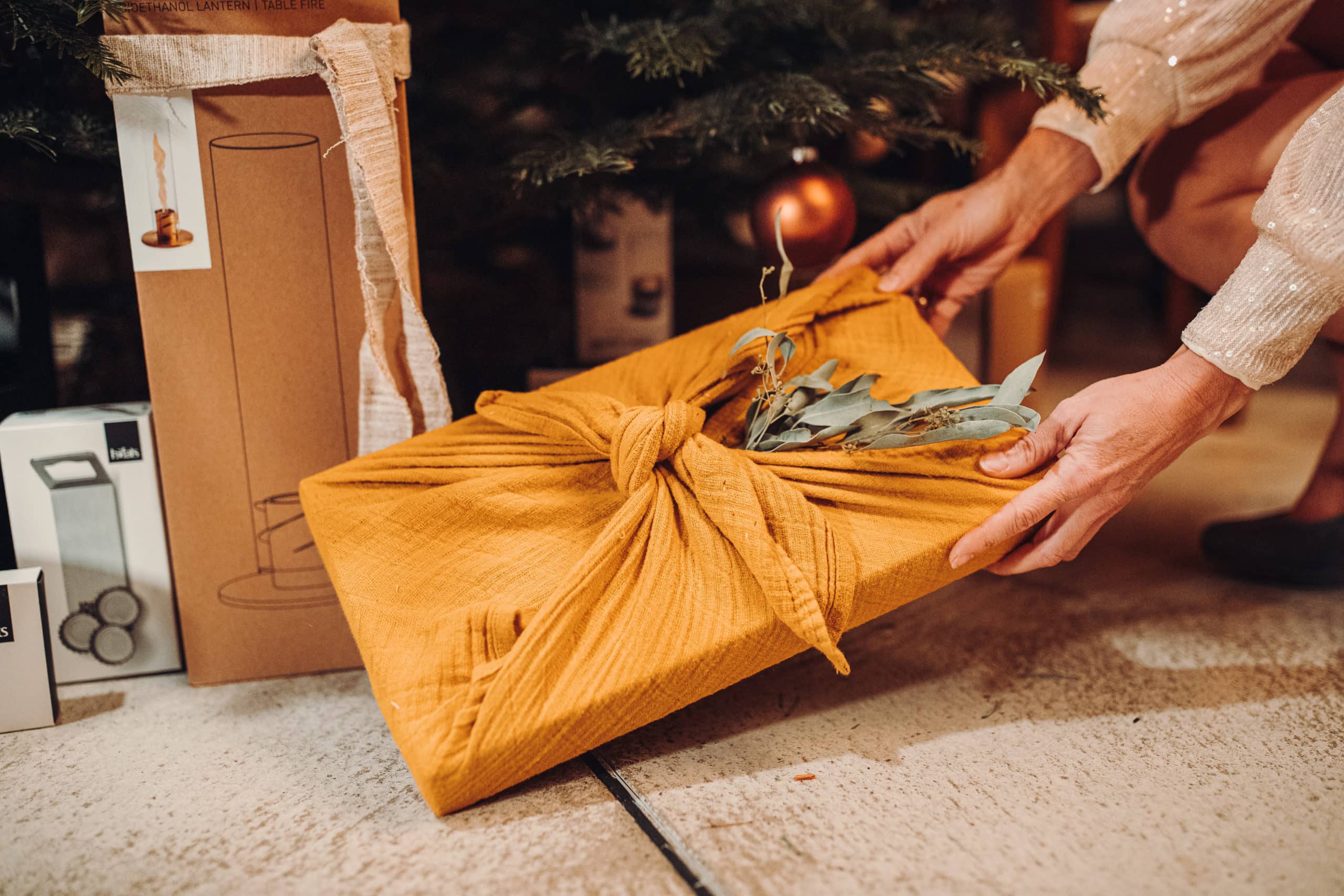 Geschenk unter Weihnachtsbaum, verpackt in gelbem Stoff mit Eukalyptus, nachhaltige Geschenkverpackung, umweltfreundlich.