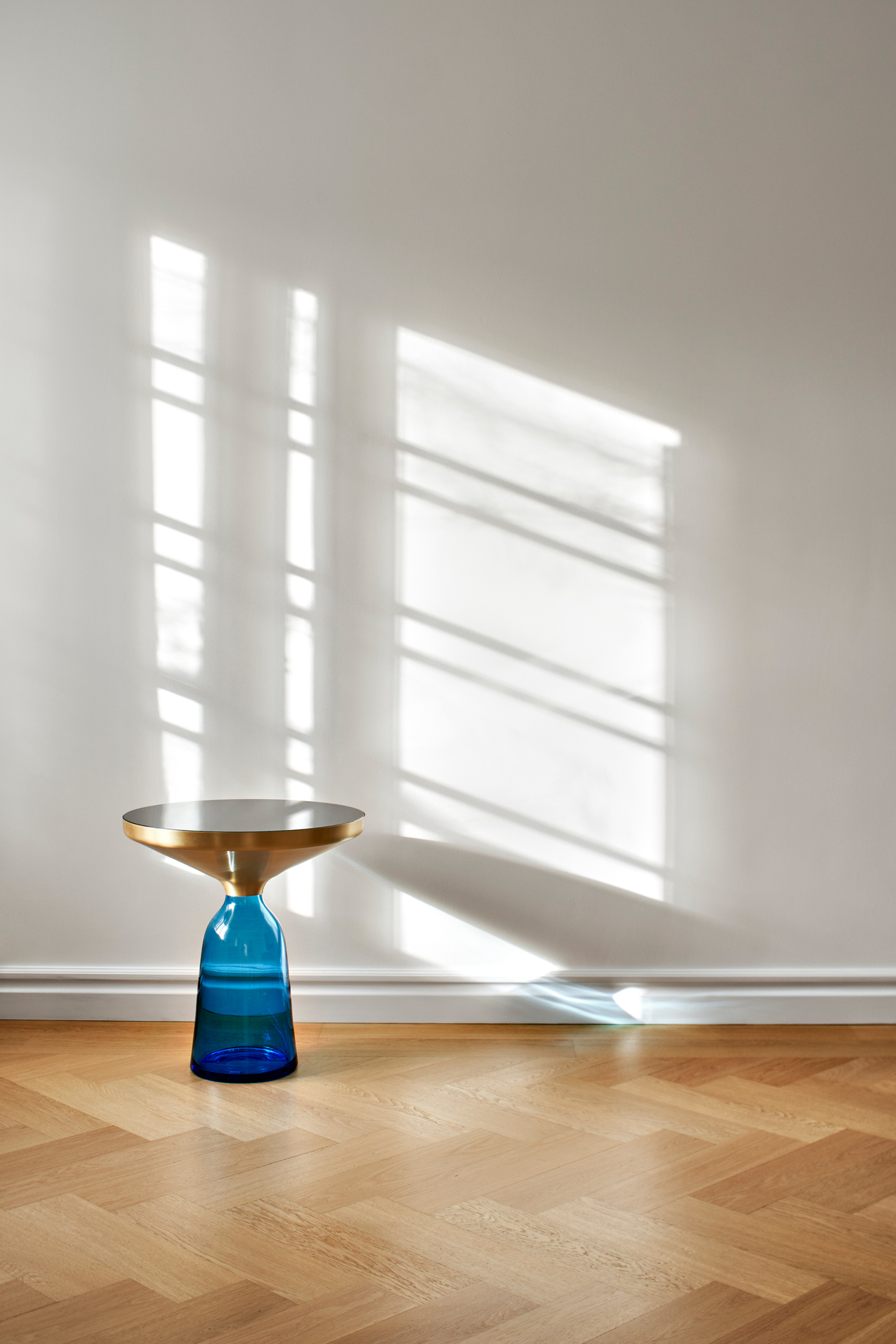 Bell Side Table Beistelltisch von ClassiCon: Blauer Glasfuß mit Messing-Tischplatte auf Parkettboden vor weißer Wand.