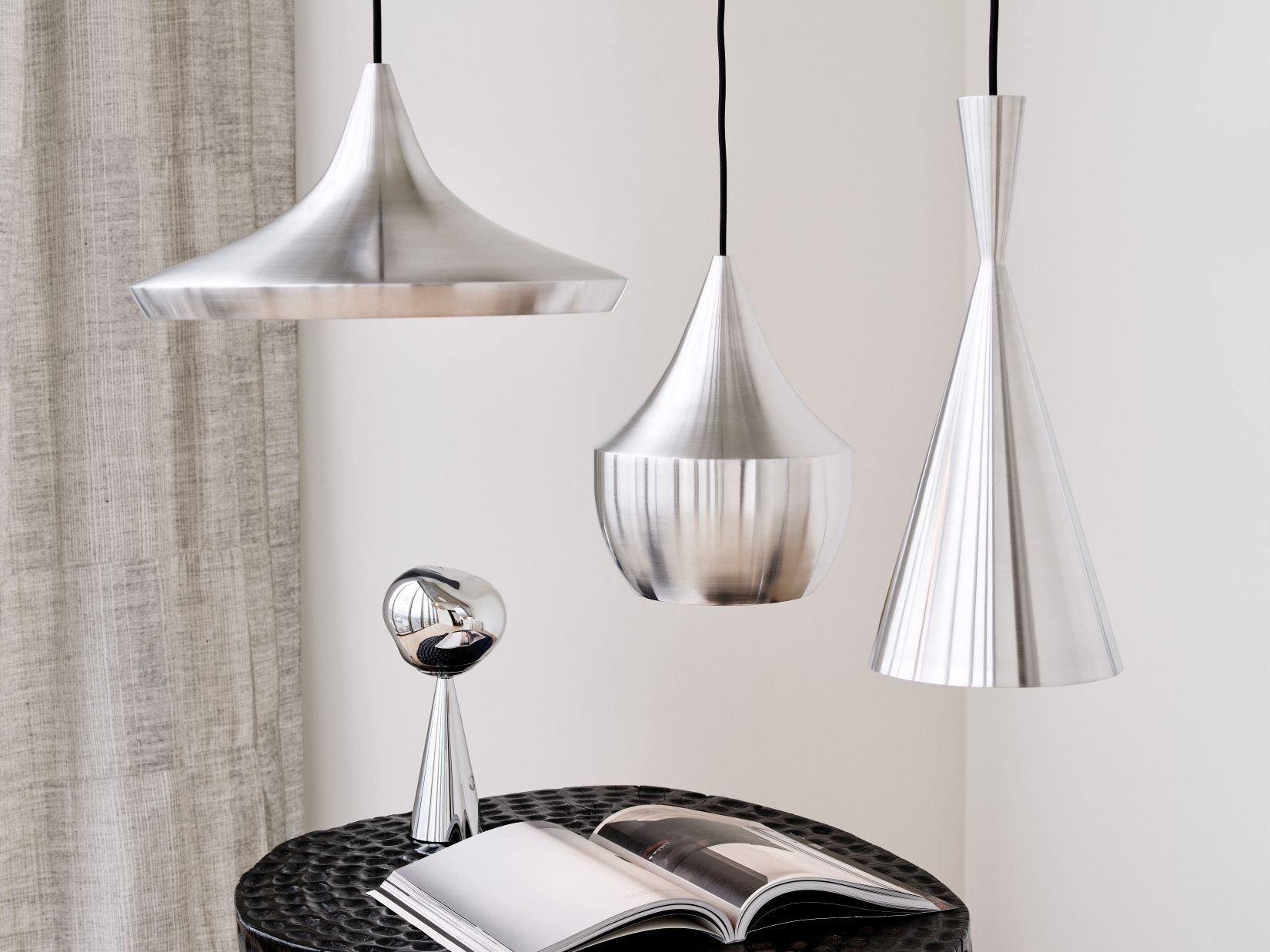 Drei silberne Tom Dixon Unbeaten Pendelleuchten in verschiedenen Formen hängen über einem Tisch mit Lampe und Buch.