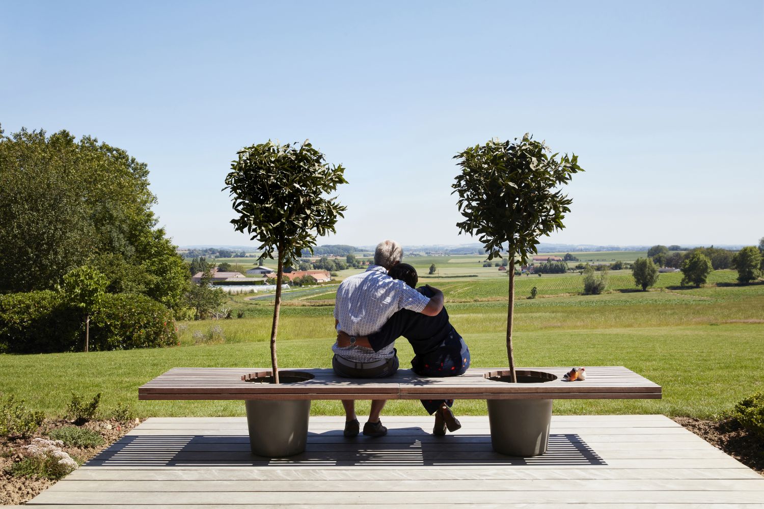 Paar sitzt auf moderner Gartenbank mit integrierten Pflanzkübeln und Bäumen, Blick auf grüne Landschaft.