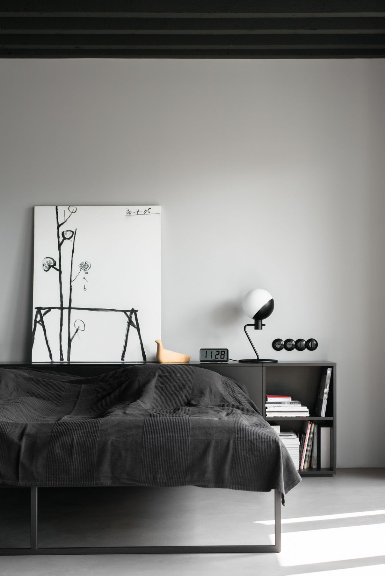 Modernes Schlafzimmer mit dunkler Tagesdecke, Nachttisch mit Lampe, Uhr und abstrakter Kunst für stilvolles Wohnen.