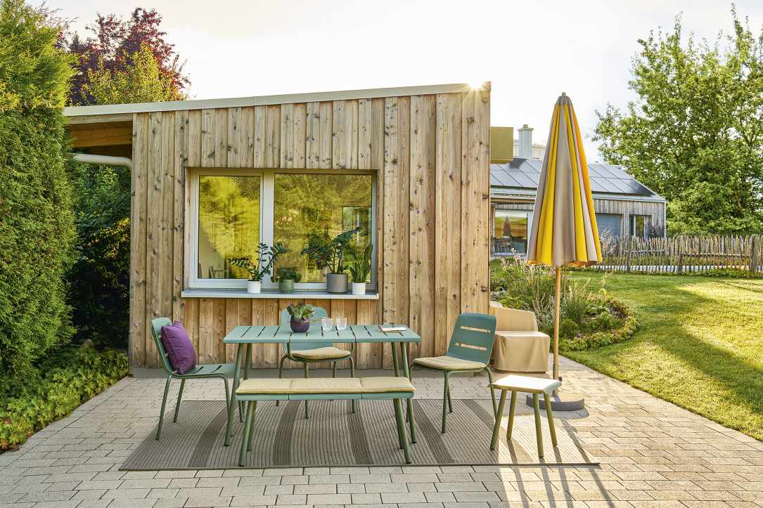 Weishäupl Vapio Outdoor Tischgruppe auf einer Terrasse mit Gartenblick, inklusive Tisch, Stühlen, Bank und Sonnenschirm.