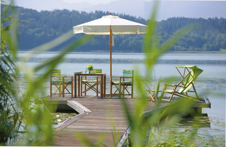 Gartengarnitur aus Holz mit Sonnenschirm und Liegestuhl auf Steg am See für entspannte Stunden.