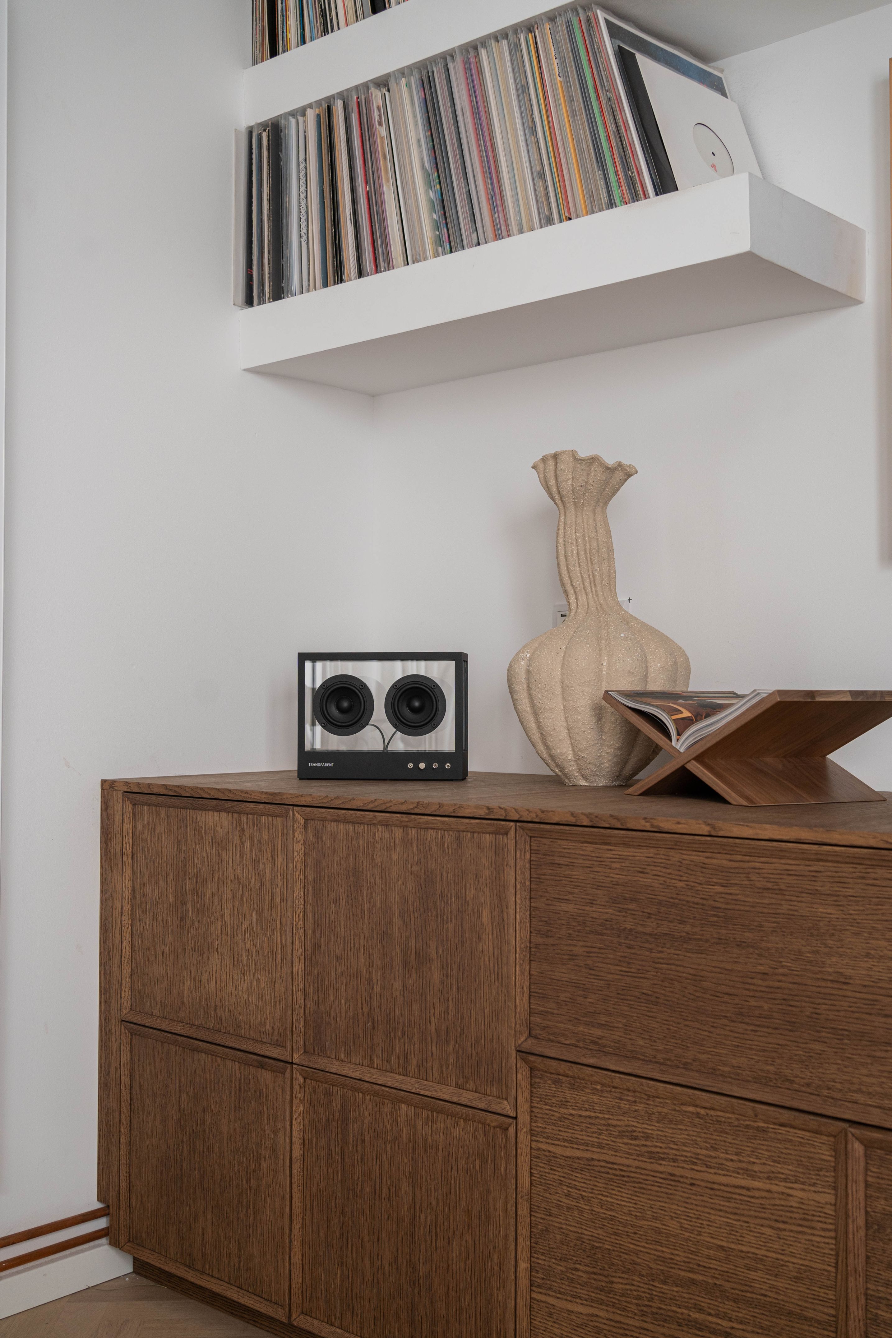 Kleiner transparenter Lautsprecher auf einem Holzschrank, daneben eine Vase und ein Schallplattenspieler mit Schallplatten im Hintergrund.