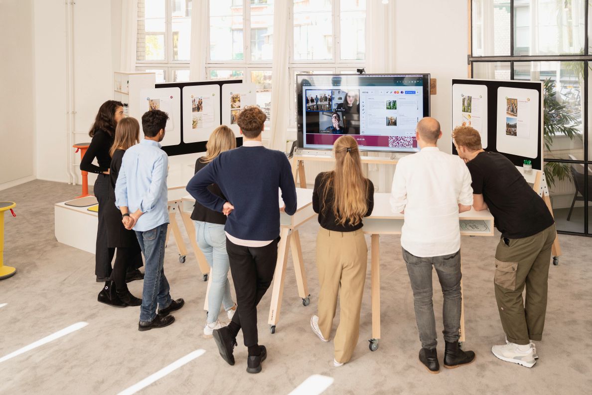 What if We Fly Taskboard: Gruppe von Personen vor Whiteboard und Medienwand im Büro.