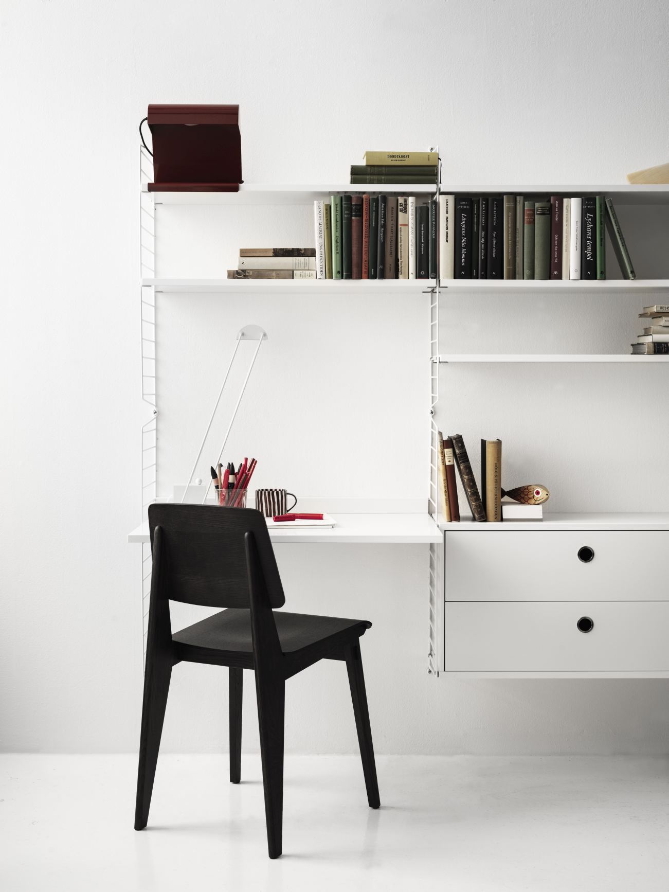 Weißes String Regal mit Schreibtisch, schwarzem Stuhl und Büchern. Arbeitsplatz-Konfiguration für Homeoffice.
