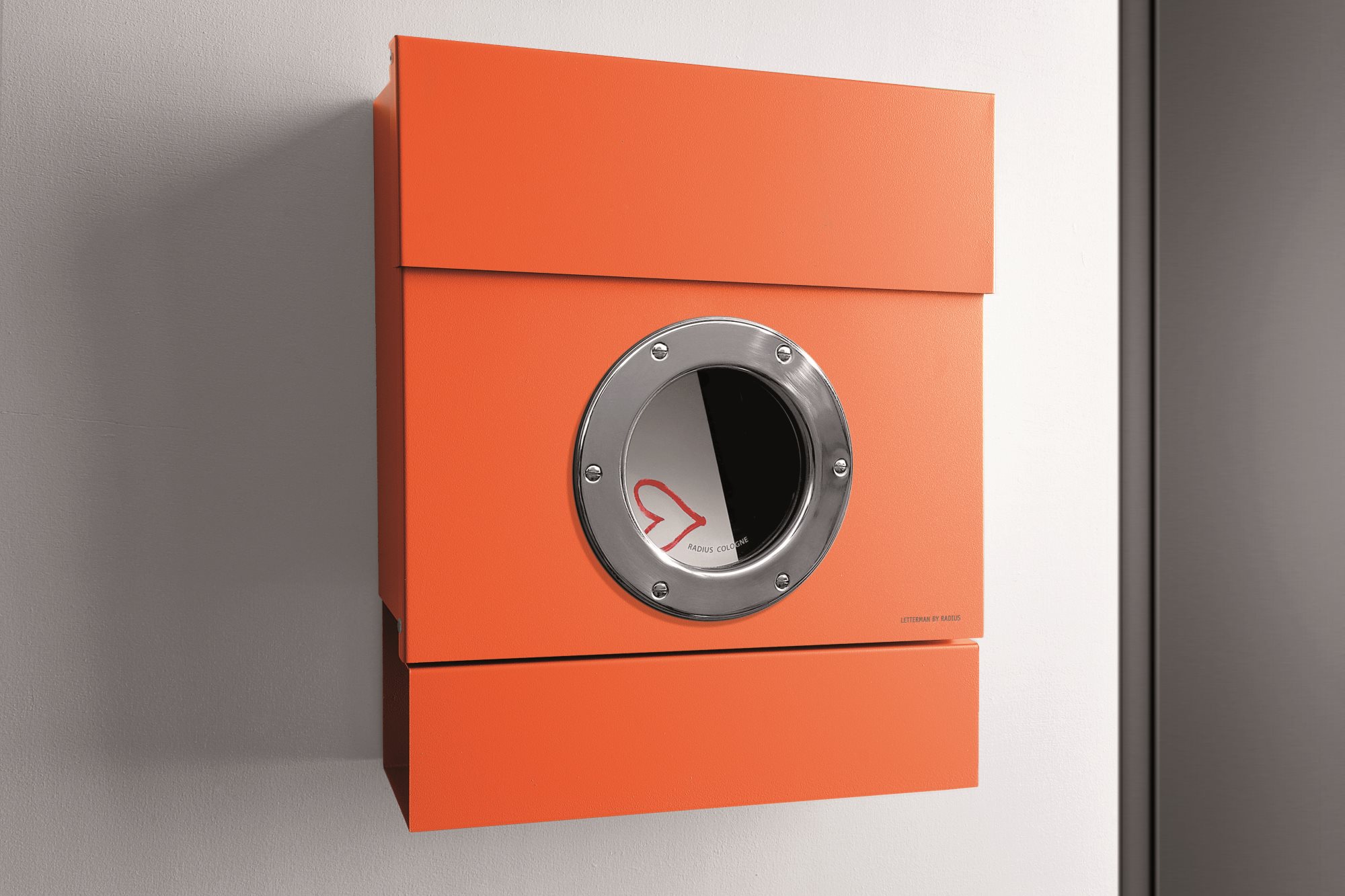 Oranger Letterman II Briefkasten von Radius mit Bullauge und Herz-Logo an einer weißen Wand.