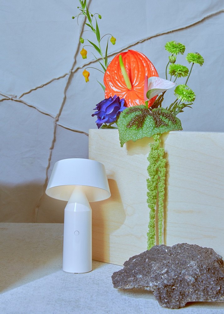 Weiße Bicoca Akkuleuchte / Tischleuchte von Marset neben Blumen und einem Stein.