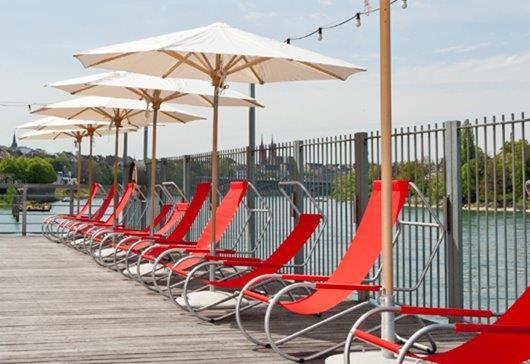 Reihe roter Lido Liegen mit Sonnenschirmen am Wasser, ideal für Entspannung und sommerliche Outdoor-Möbel.