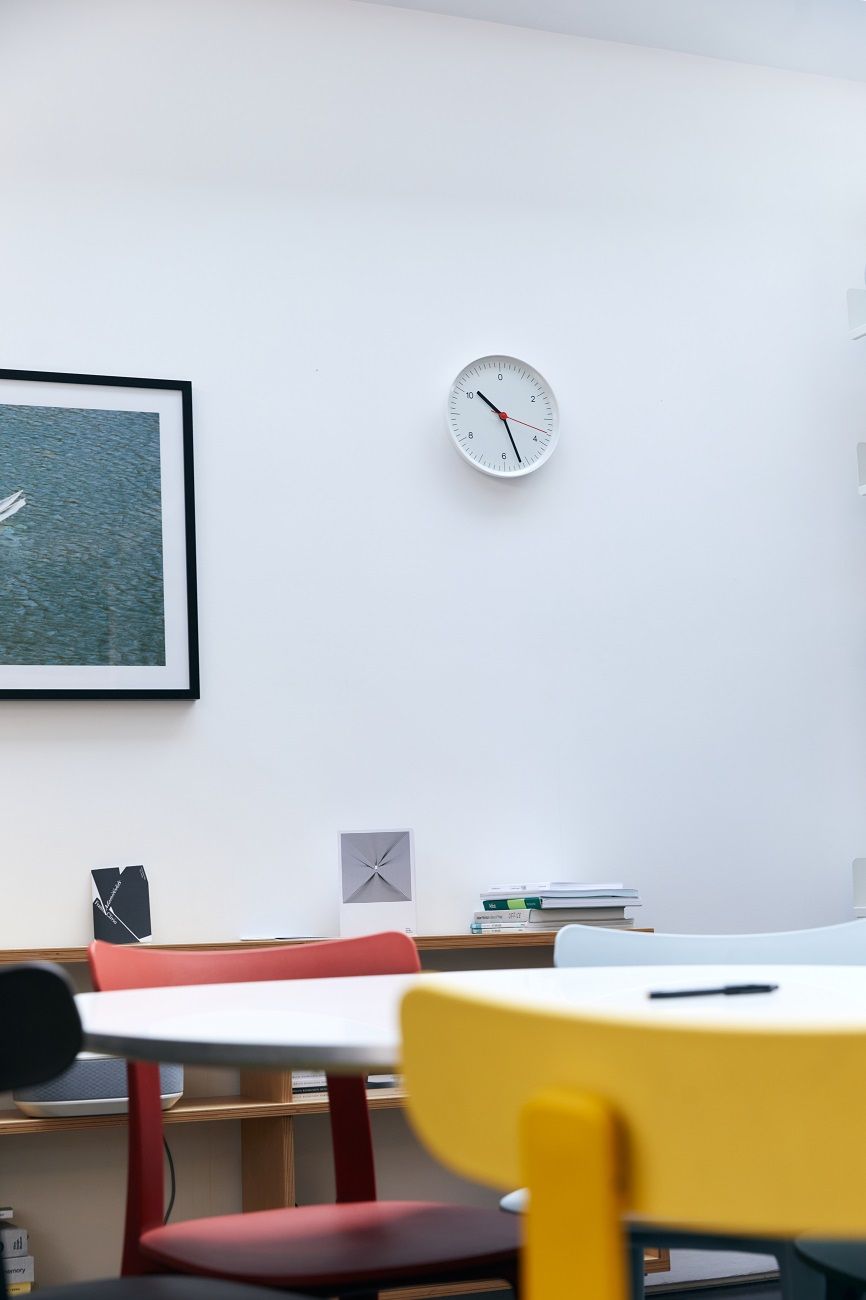 Weiße Hay Wanduhr mit minimalistischem Design an einer weißen Wand über farbigen Stühlen.