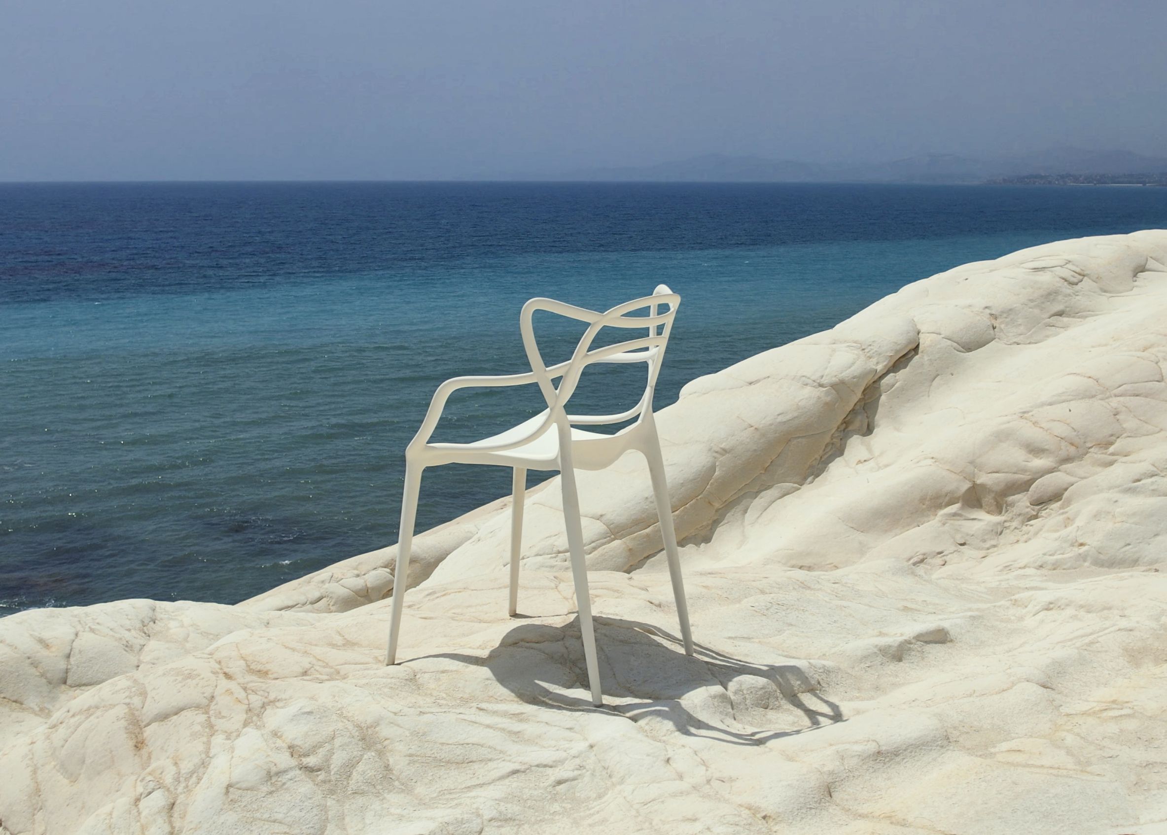 Weißer Masters Stuhl von Kartell vor blauem Meer und hellem Felsen. Designstuhl für Garten.