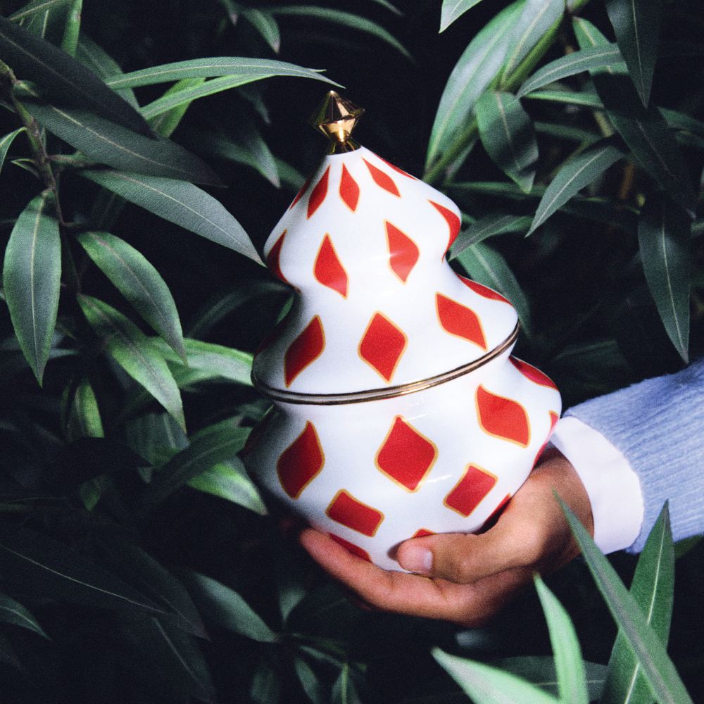 Alessi NZ03 Delight Behälter für Süßigkeiten, gehalten in der Hand, weiß mit roten Rauten und goldener Spitze.