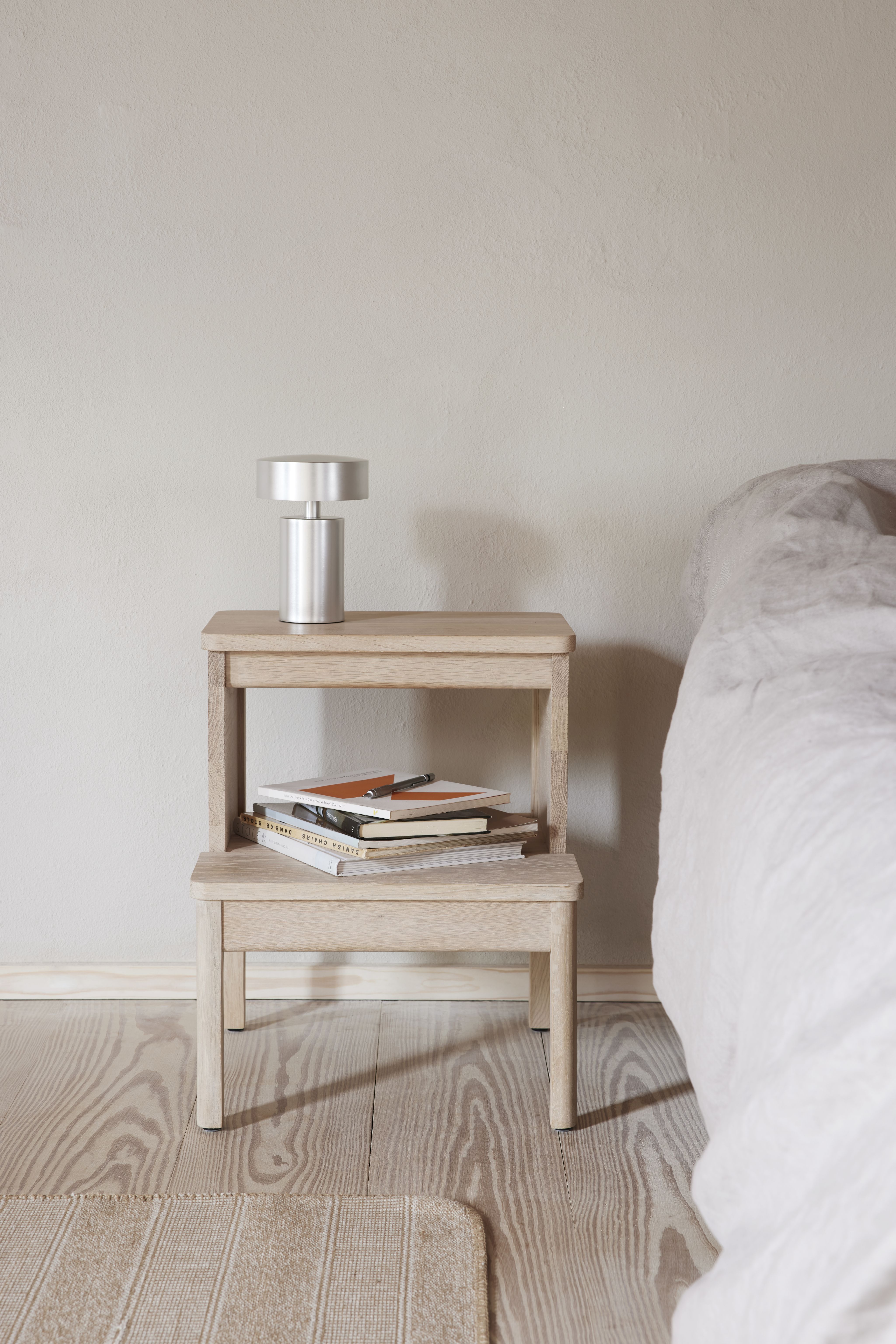 A Line Steighocker von Form & Refine aus hellem Holz als Nachttisch mit Lampe.
