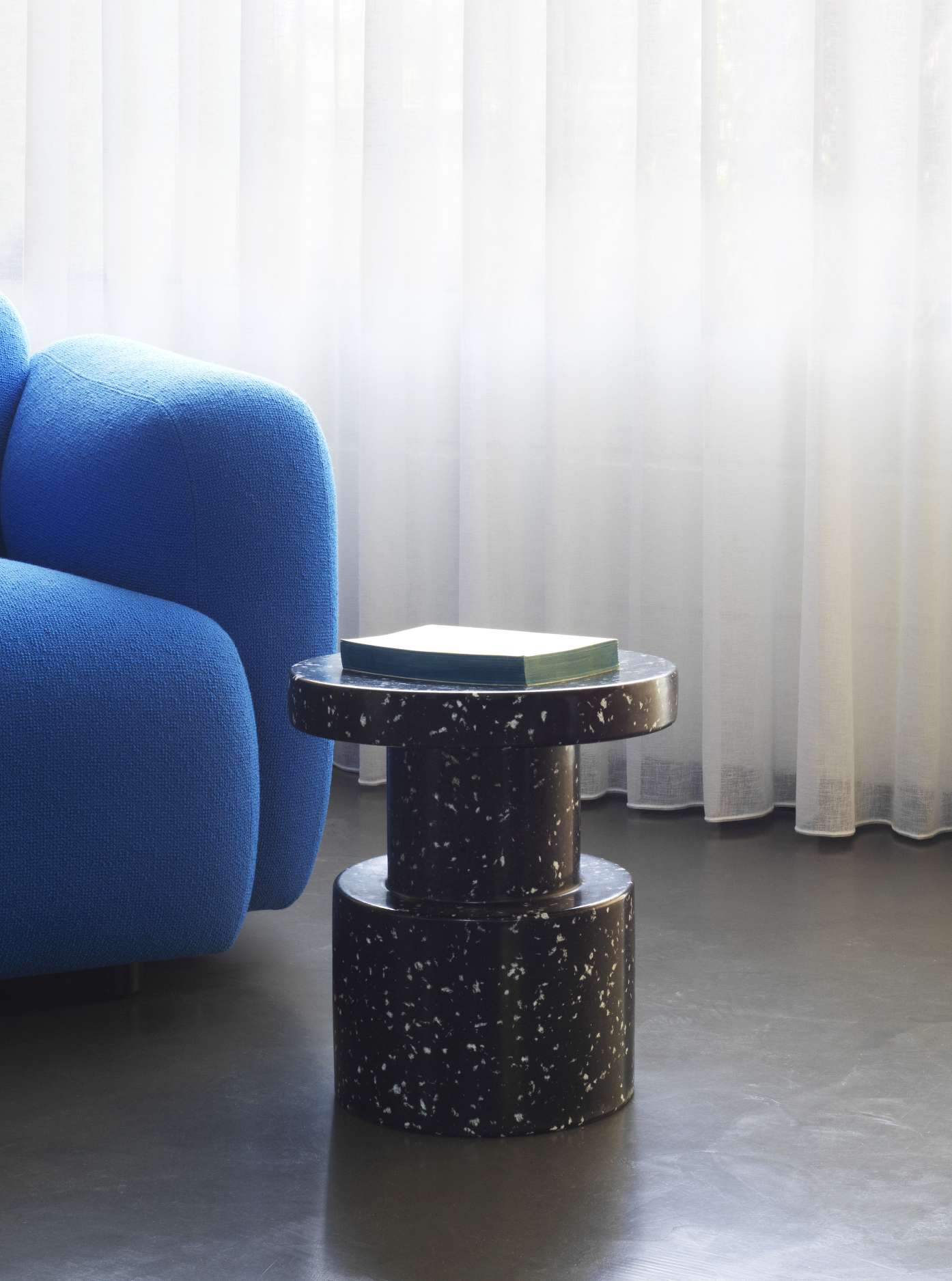 Normann Copenhagen Bit Stool Hocker in Schwarz-Weiß-Optik neben einem blauen Sessel und Vorhängen.