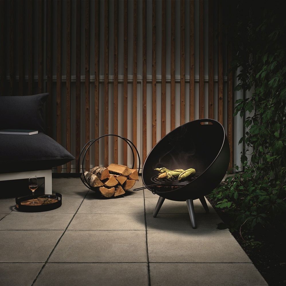 FireGlobe Brennholzhalter neben Grillstelle mit Maiskolben und Holzscheiten im Garten.